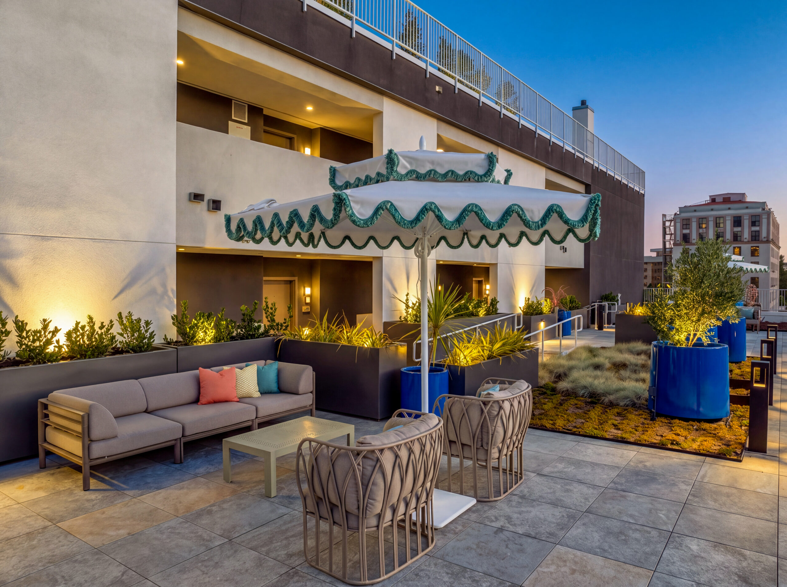 A rooftop patio at twilight with couches, chairs, umbrellas, and a view of Downtown Los Angeles.